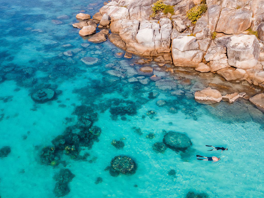 Aerial view of two people snorkeling in crystal Blue water and coral reef near a rocky island.