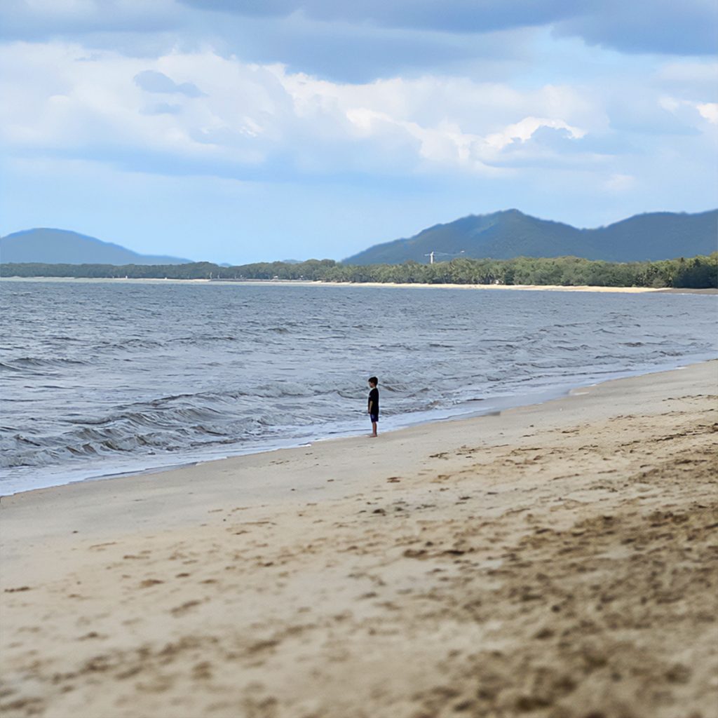 Boy standing at the edge of the shoreline of a beach at Palm Cove. Mountains and forest in the background.